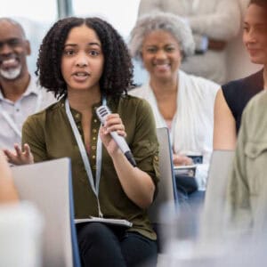 istockphoto-1134043640-612x612 Young African American woman gestures while asking a question during a seminar or conference.